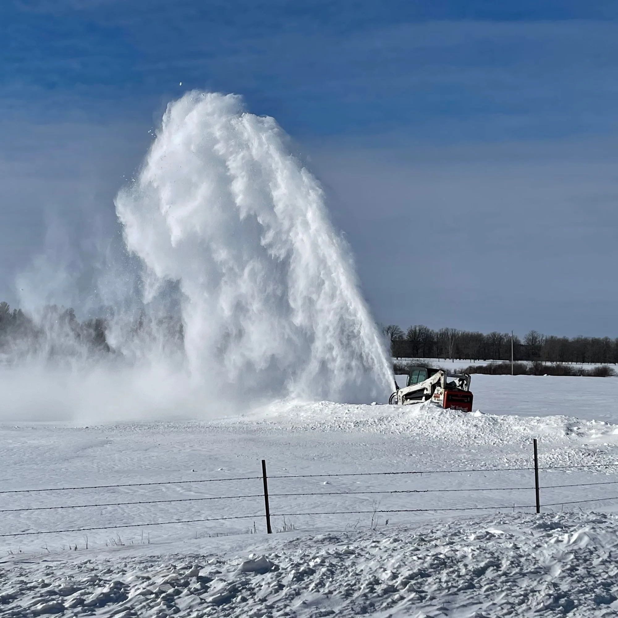 Shepard's skid steer blowing a tall plume of snow across a snow-covered property